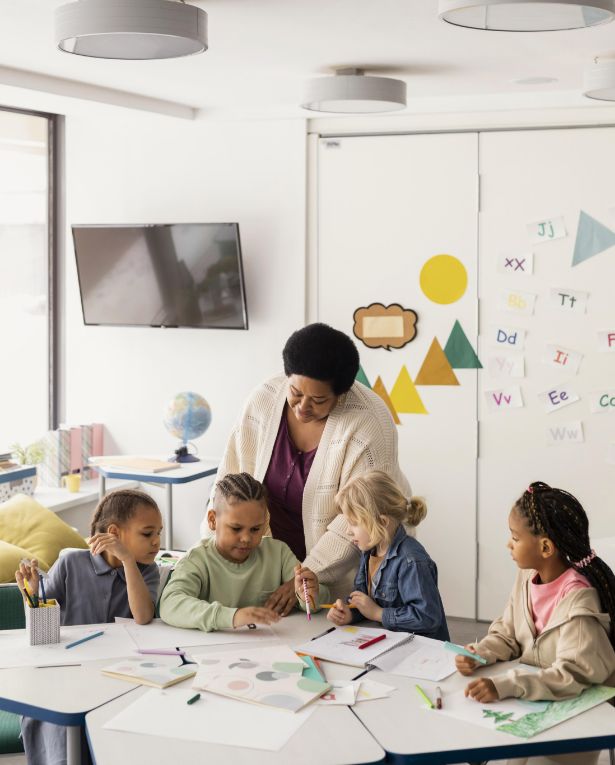 A teacher helps four young children with a drawing activity at a classroom table. The kids are focused on their work, surrounded by colored pencils and paper. The classroom has bright decor and educational posters on the wall.