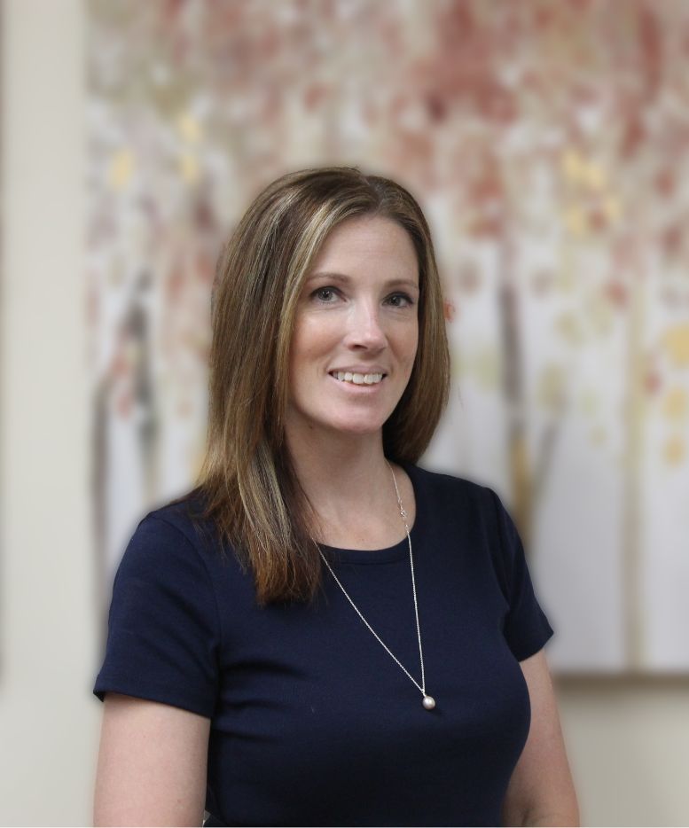 A woman with straight, shoulder-length brown hair wearing a navy blue top and a pendant necklace stands in front of a blurred background with abstract art featuring soft, warm colors. She is smiling slightly.
