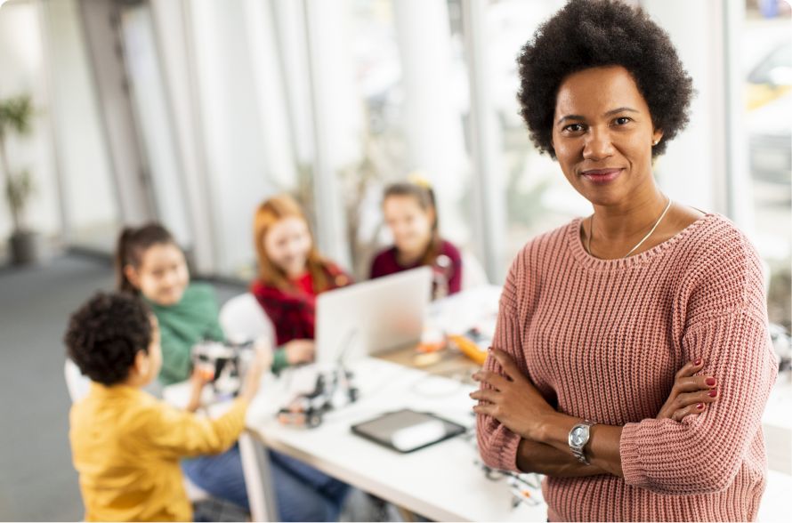 A confident woman stands with arms crossed and smiling in the foreground, while four children work together on a robotics project at a table in the background.