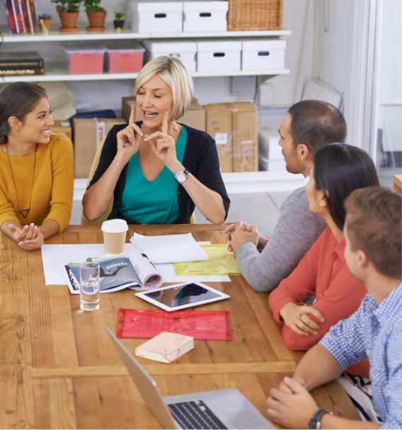 Five people sit around a wooden table in a bright office, engaged in discussion. Papers, a laptop, and a tablet are on the table. One woman is speaking animatedly while others listen and smile.