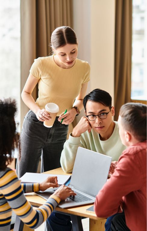 Four young adults gather around a table with a laptop, engaged in discussion. One woman stands holding a coffee cup, while the others sit, listening and talking in a bright, casual setting.