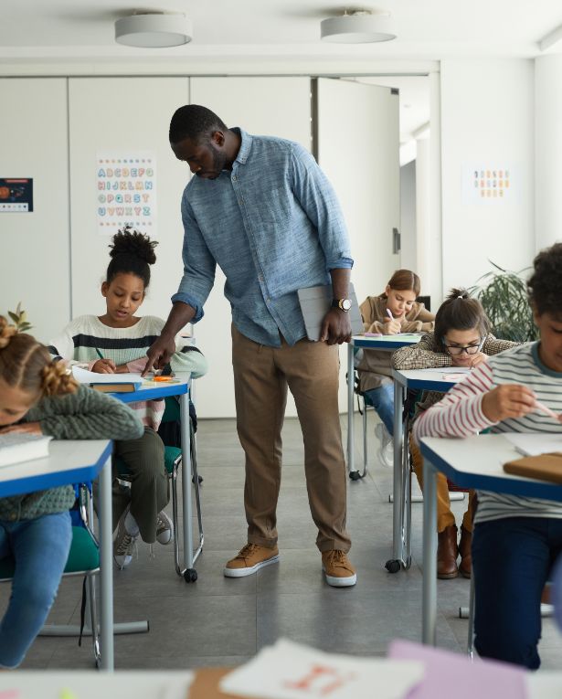 A teacher stands by a students desk, assisting her, while other students sit at tables working on assignments in a bright classroom.