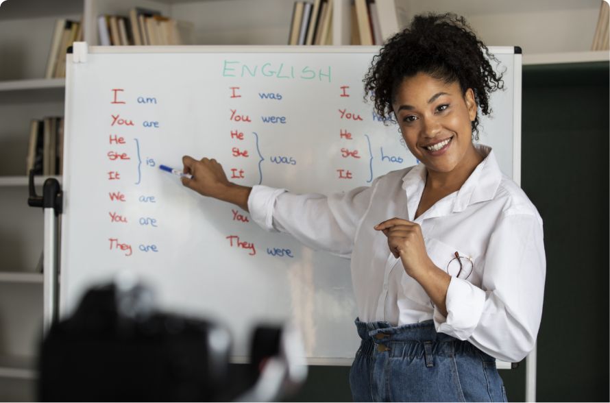 A woman stands in front of a whiteboard teaching English grammar, pointing to pronouns and verb forms. She is smiling and wearing a white shirt, with bookshelves in the background.