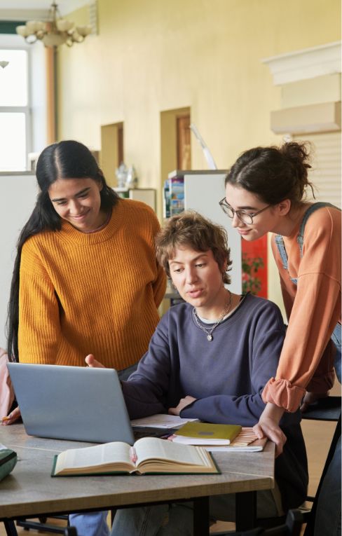 Three young adults gather around a table, looking at a laptop screen and discussing something. Open books and notebooks are on the table, suggesting a study or group work setting.